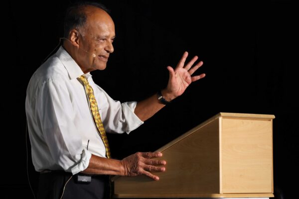 Professor Sir Partha Dasgupta stands at a lecturn. He raises his hand while he is speaking.