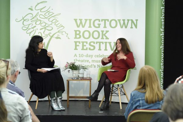 Gerda Stevenson sitting on stage with chairperson Lee Randall at Wigtown Book Festival. A table containing flowers, water and glasses is in between them. The audience sit before them listening.