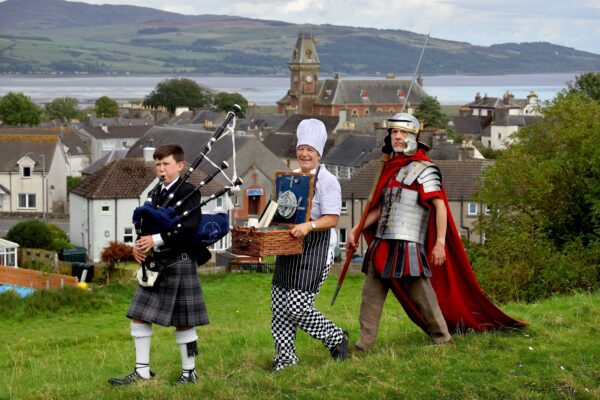 One piper piping, one chef carrying a picnic basket and one centurion marching, on a hill with Wigtown and Wigtown bay behind them.