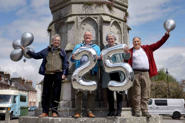 Four people standing in front of  Mercat Cross, Wigtown, holding silver balloons for the twenty fifth anniversary of Wigtown's Book Festival.