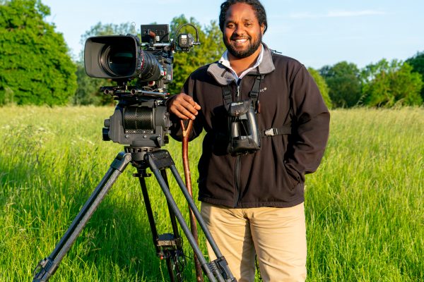 Hamza Yassin is standing in a grassy field with a large camera on a tripod. Blue skies with wispy clouds.