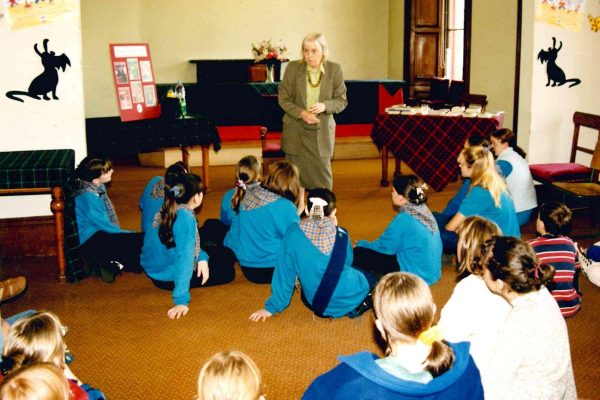 Joan Linguard is standing in a room talking to a group of GirlGuides. They are sitting on the floor in Wigtown County Buildings.. A table is covered with a tartan tablecloth and books.