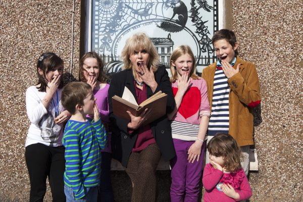 Actress Joanna Lumley standing holding an open book surrounded by six children in Wigtown, Scotland's National Book Town. They all have their hands to their mouths and are gasping. Behind them a pebble dashed wall and a window with a black and white etched picture featuring a house, a fox, a bird, flowers and a large cobweb in the left hand top corner.