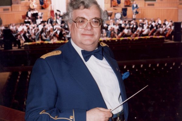 John Mason stands in formal attire holding a baton, the Scottish Fiddle Orchestra behind him.