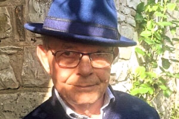 Author John Wheway head and shoulder portrait standing in sunshine wearing blue hat