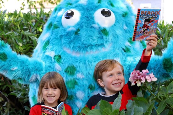 Two children in red and black striped Dennis the Menace tops hold Beano books. Big Wig, the big blue mascot, stands behind them with arms outstretched. They are in a garden surrounded by shrubbery.