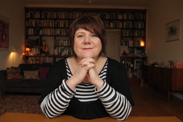 Louise Welsh is sitting at a desk in a room with bookshelves on the wall, a settee, sideboard, pictures and lamps behind her.