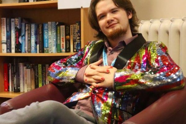 Thomas McClure is sitting in an armchair beside a bookshelf full of books. He is wearing a colourful jacket and has his hands clasped together.
