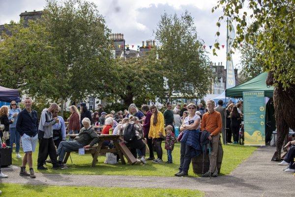 People gathered on a sunny day in Wigtown town gardens during Wigtown Book Festival. A pop up cocktail bar is serving drinks, people are eating at the picnic benches.