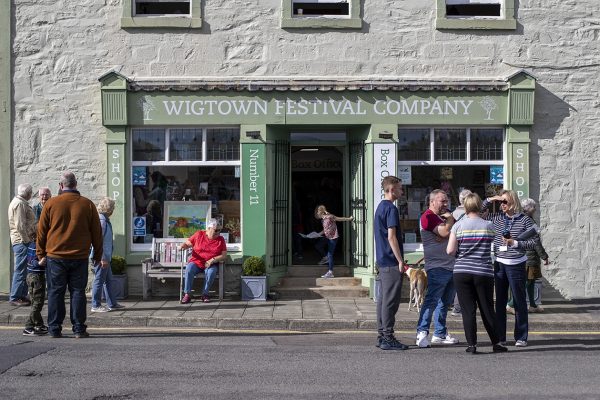 Many people are standing outside Wigtown Festival Company bookshop and box office talking and enjoying the sunshine, during Wigtown Book Festival.