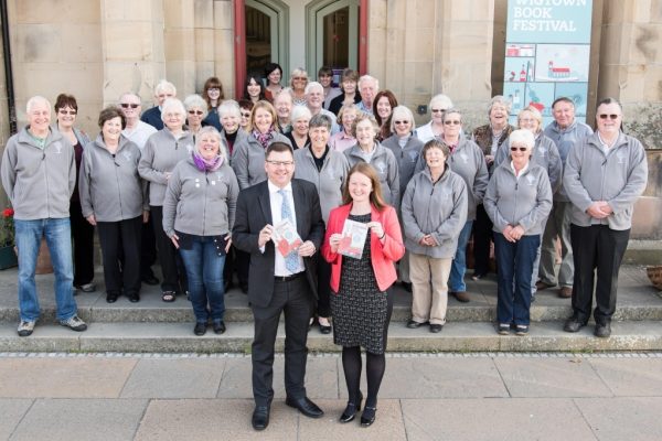 A large group of Wigtown Book Festival volunteers gathered on the County Building steps smiling. Anne Barclay and colleague hold the program brochure.
