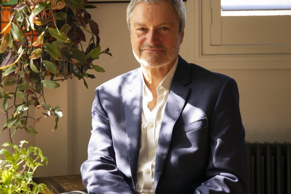 Gavin Esler sitting in a room. Windows with slatted blinds and a large hanging plant beside him.