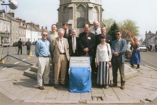 Group of men and one woman standing in front of Mercat Cross, Wigtown, unveiling a plaque to commemorate the launch of Scotland's national Book Town. In the background bookshops, cars and people with one women dancing merrily.