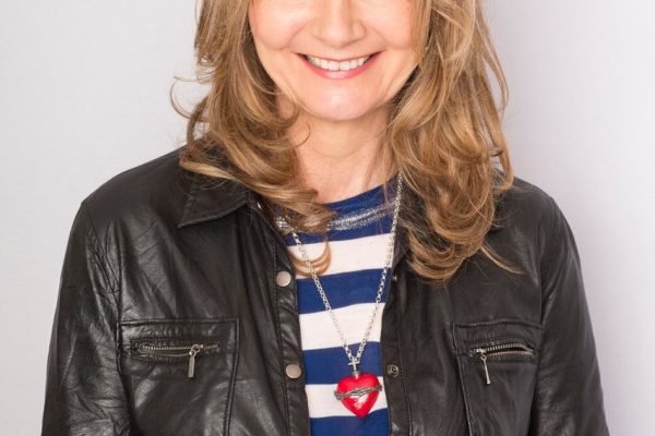 Author jo Caulfield stands smiling wearing a black jacket and blue and white striped t-shirt.