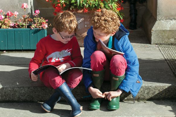 Two young children sitting on steps looking at a book together in the sunshine at Wigtown Book Festival. Colourful flowers are in pots behind them.