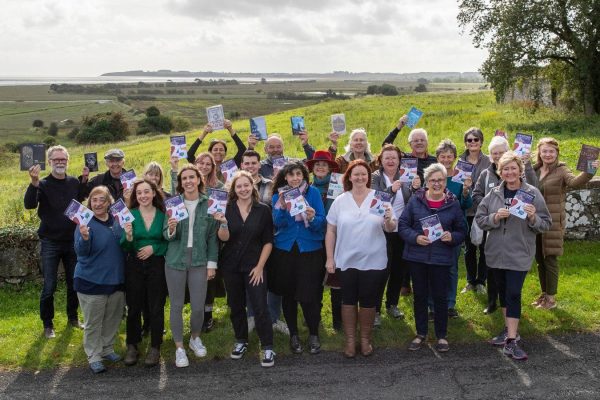 Group picture of Wigtown Book Festival staff and volunteers standing in front of Wigtown Bay smiling and holding up Festival brochures.