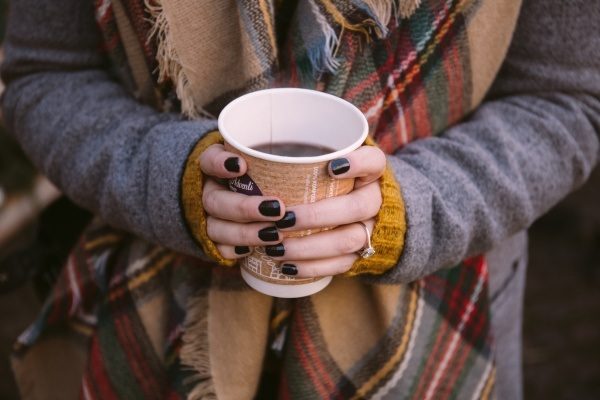 A person wearing a tartan scarf is holding a cup of mulled wine. Their fingernails are painted dark crimson.