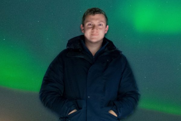 Nathan Case is standing in a heavy coat with the Northern Lights displayed behind him.