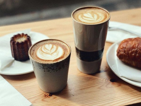 On a table stand two cups of coffee latte in brown pottery, a pastry and a small cake on plates.