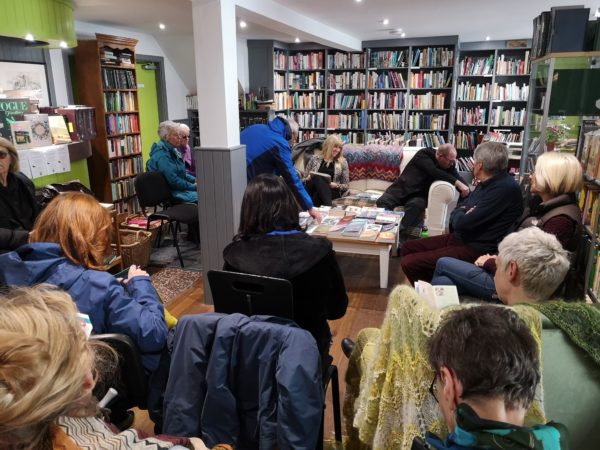 A room full of books on bookshelves. People are sitting on chairs and a couch talking and smiling. A table is covered in books in the center of the picture.