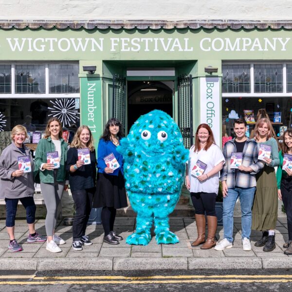 Eight staff members stand either side of Big Wig, the big blue book festival mascot, outside the number 11 bookshop. Each staff member is holding a 2023 festival brochure in front of them.