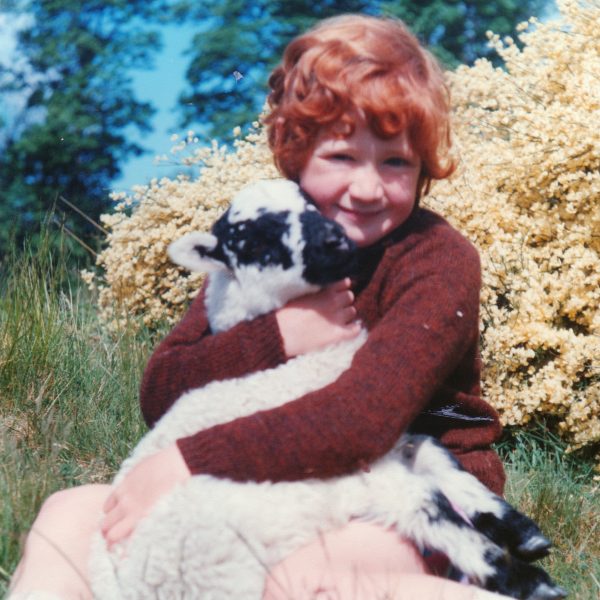 Old photo of Polly Pullar as a child. She is sitting in a small field in front of a yellow flowering bush holding a lamb in her arms.