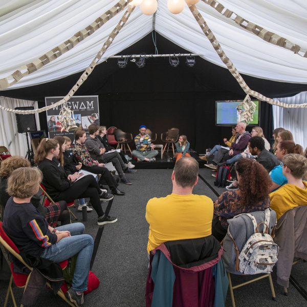 A mixed group of people are sitting in a circle at an event at Wigtown Book Festival. They are listening to a speaker sitting crossed legged on the floor. The ceiling is draped with white material and garlands are strung from the lights above them.