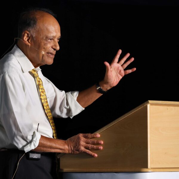 Professor Sir Partha Dasgupta stands at a lecturn. He raises his hand while he is speaking.