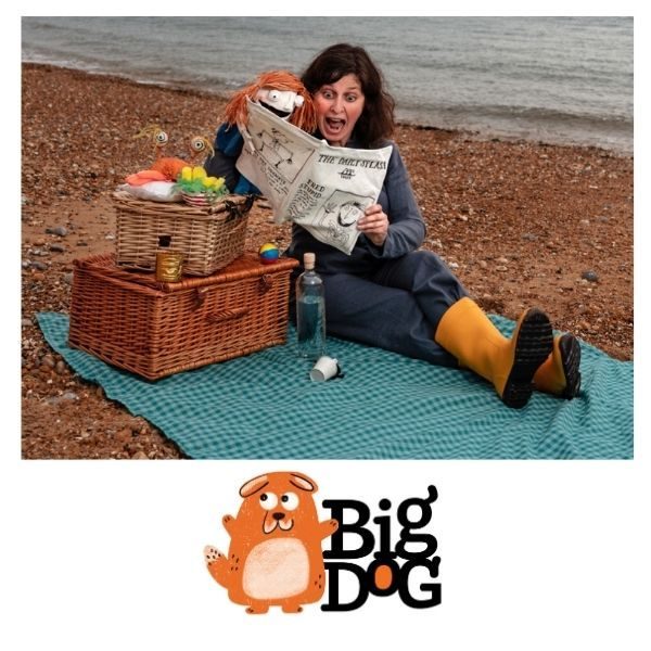 Jo is sitting at the beach on a blanket with two picnic baskets. She and a hand puppet are reading a book. A Big Dog Children's Book Festival event.