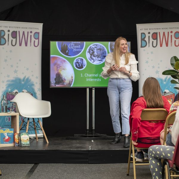 An author stands on the stage at her Children's Book Festival event. She is talking animatedly to a young audience sitting on chairs in front of her. Books and a poster are on the stage with her.