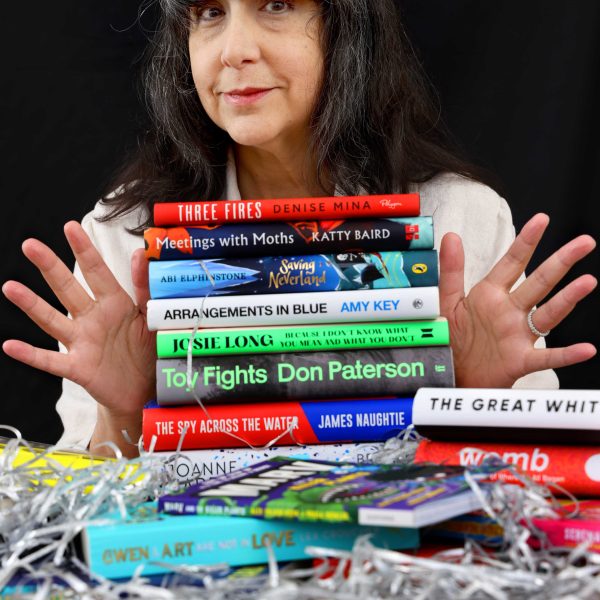 Lee Randall, Guest Programmer for Wigtown Festival Company stands behind a stack of books holding her hands out to either side of the books.