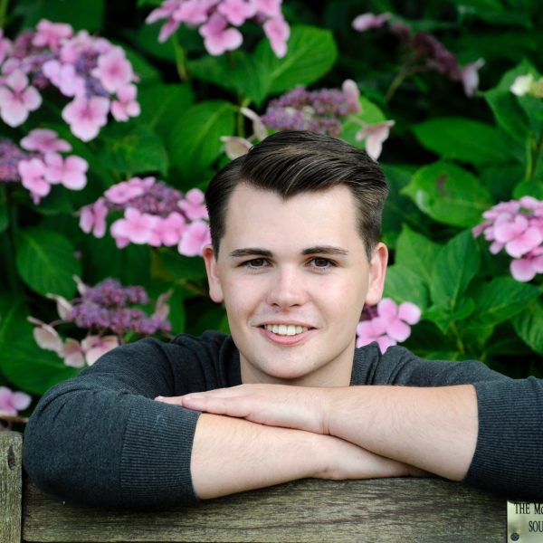 Andy Stewart, Children and young people's producer,  stands in front of a pink hydrangea bush in Wigtown gardens with his arms folded in front of him.