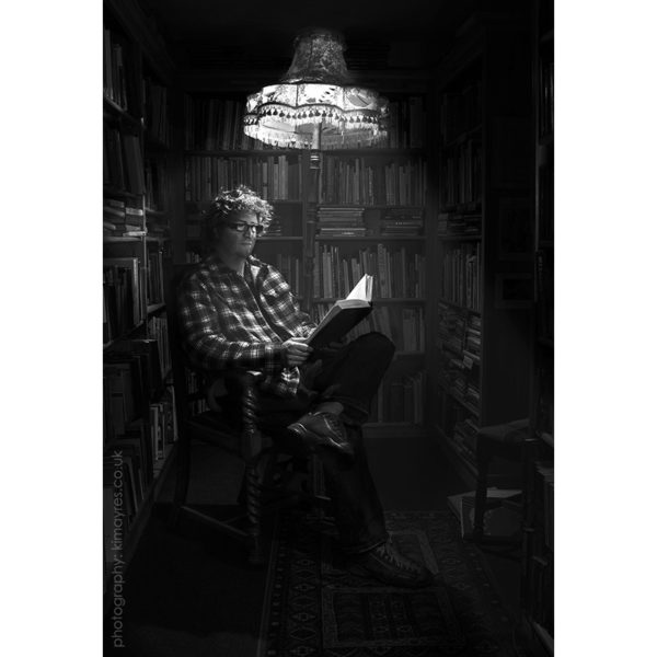 Black and white photo of Shaun Bythell sitting in a chair reading a book under the glow of a tall lamp. Bookshelves full of old books surround him.