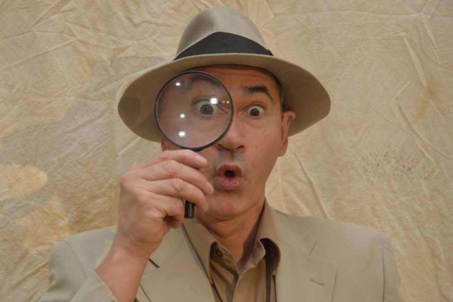 Man smartly dressed in a suit and hat holding a magnifying glass to his eye, with a shocked expression on his face.