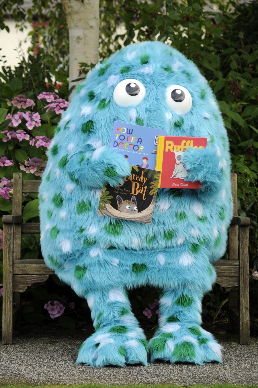 Bigwig, the Children's Book Festival mascot, stands in front of a bench in Wigtown Gardens holding books.