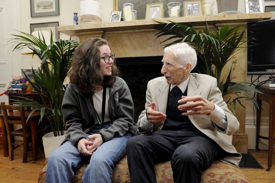 Lord Martin Rees is sitting talking animatedly to Helena Cochrane. A large stone fireplace with photo frames and candles is behind them. Potted plants, a television and a desk with a chair are either side of the fireplace.