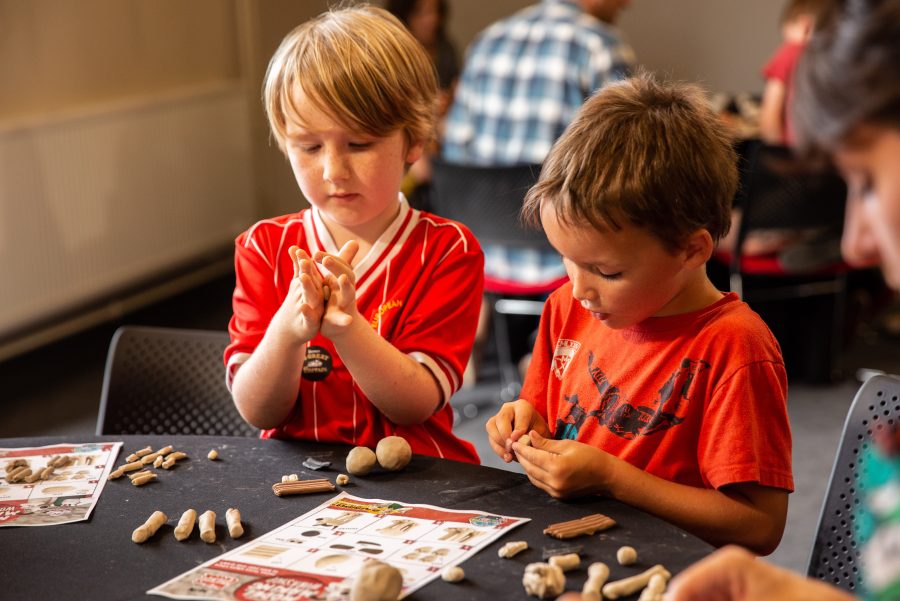 Two young children making models out of modelling clay during a Children's Book Festival event. While sitting at a table they are rolling out small balls of clay, following instructions from a sheet of paper.