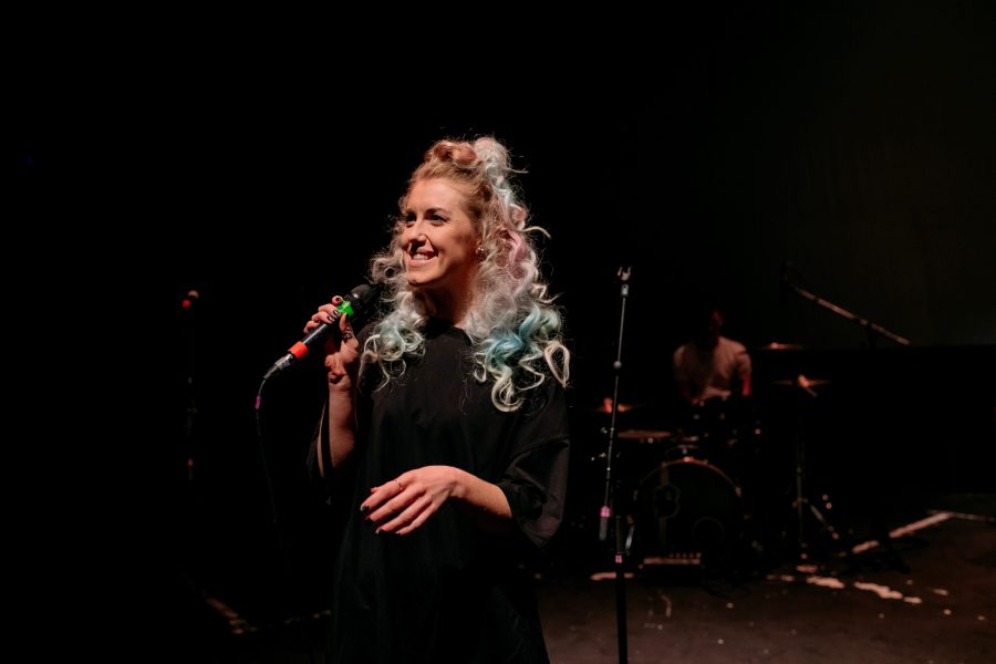 Imogen Stirling is standing singing into a microphone during a music event. A drum set and drummer are behind her.