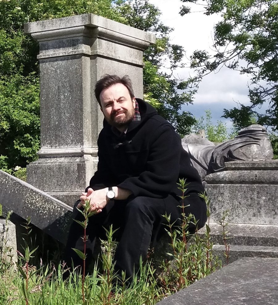 Peter Ross crouching down beside an old stone pillar in a church graveyard.