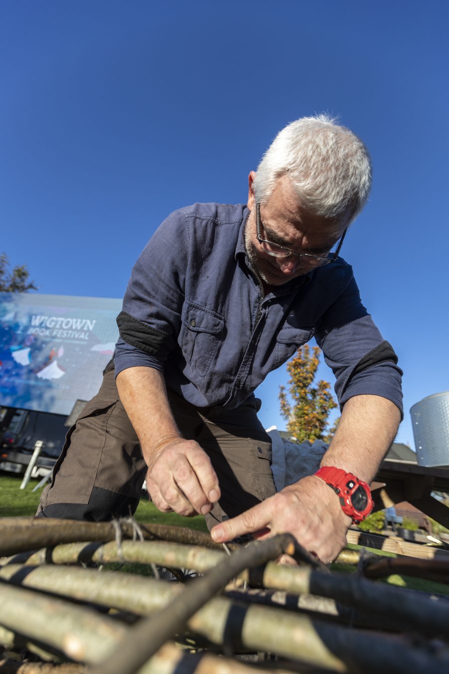 Robert Twigger is kneeling down building his coracle. A small one-person boat made out of woven wood and a waterproof covering. Behind him the large screen showing Wigtown Book Festival events.