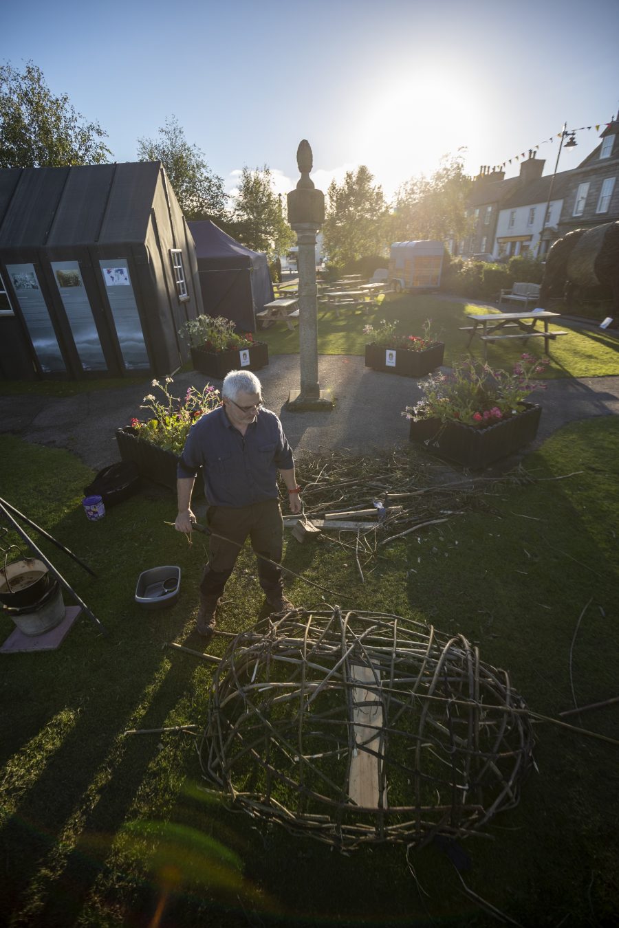 An overview of Robert Twigger building his coracle. A small wooden woven boat. He is standing in the gardens at the Wigtown Book Festival. The Deception Island Hut stands behind him.