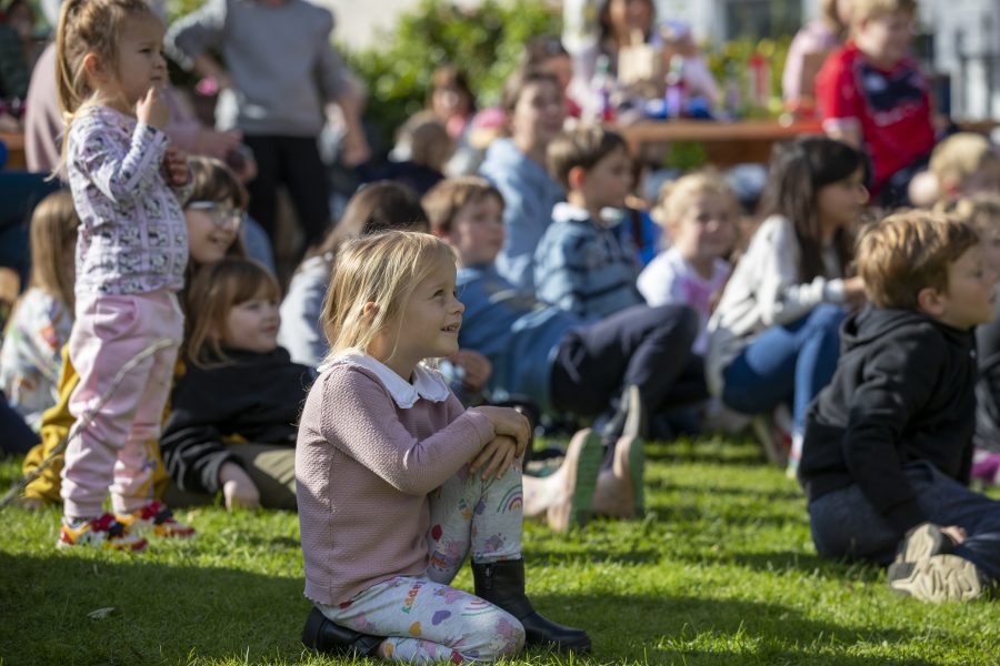 Many young children sit listening at an outdoor Children's Book Festival Event in Wigtown gardens.