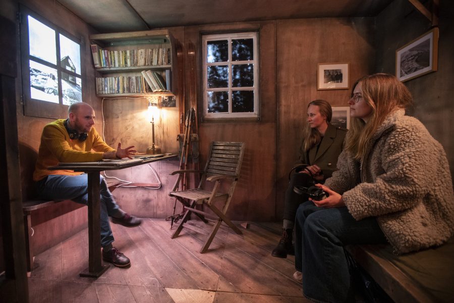 A speaker and two people are sitting inside a replica refuge hut to glimpse what life was like for members of the British Antarctic Survey based in one of the most remote places on the planet. Inside the hut there is a table with a lamp a chair and a bench, old walking poles are standing up against the wall. A screen on the wall is showing images.