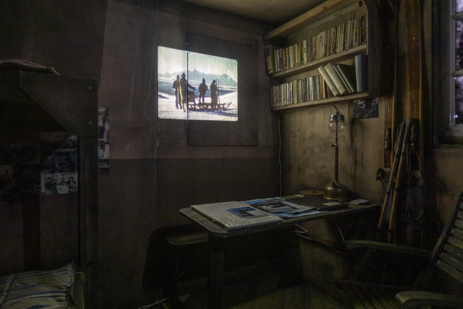 Inside the Deception Island Hut at the Wigtown Book Festival. A table with a lamp, books and maps, a small bookshelf with old books on the left. Bunkbeds are on the right side of the hut. A slideshow is playing on the wall.