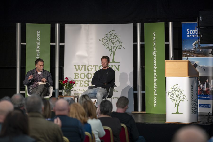 Darren McGarvey sitting on stage with a chairperson for his event at Wigtown Book Festival. The audience are sitting in front listening to them. Various banners adorn the stage, a small table with flowers and water glasses sits between the men.