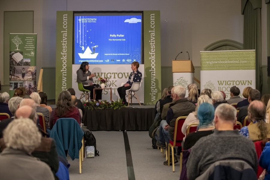 Polly Pullar and Lee Randall sit on stage talking at her event during Wigtown Book Festival. The audience sit in rows in front of them listening. At the back of the stage a large screen is showing the author's name and book title.