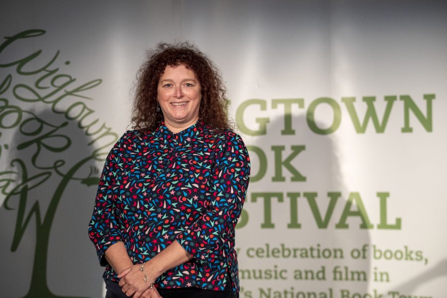 Wigtown Book Festival Poetry Prize winner 2022 Julie Laing. She stands in front of the green and white Wigtown Book Festival logo.