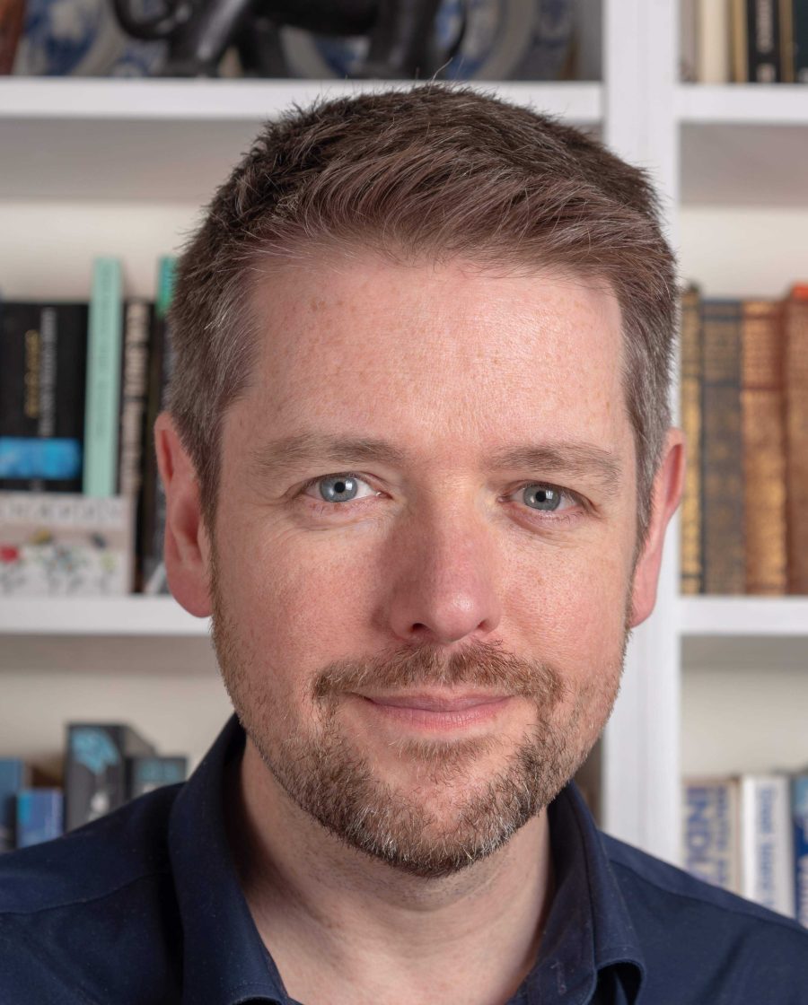 Headshot of Alastair Chisholm. Bookshelves containing books behind him.