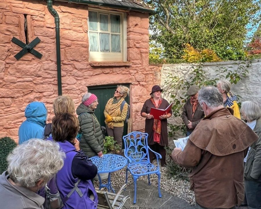 People standing in the small back yard of a sandstone house, listening to a speaker during a Dorothy L Sayers walk for Wigtown Book Festival.