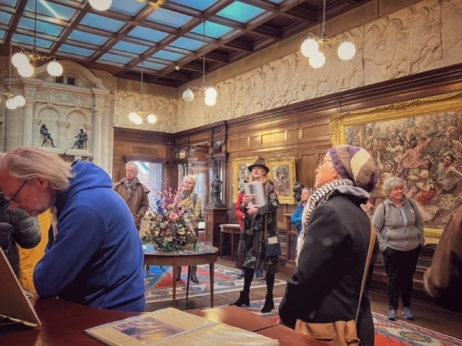 Many people stand listening inside an ornate room to a speaker during a Dorothy L Sayers walk event for Wigtown Book Festival. Large oil paintings adorn the walls, bright rugs on the floor.